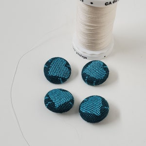 May include: A spool of off-white thread and four teal and black fabric-covered buttons are arranged on a white surface. The thread spool has text that reads "Gütermann Germany". The buttons have a patterned design.