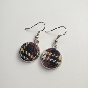 May include: A pair of round button earrings with a plaid pattern in black, brown, and cream colors. The earrings have silver-colored hooks and a silver-colored metal frame around the button.