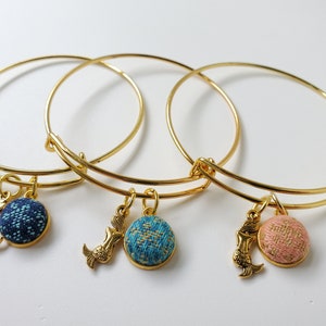 May include: Three gold-colored bangle bracelets. Each bracelet has a round charm with a textured fabric design in blue, teal, and pink. A gold mermaid charm hangs next to each fabric charm. The bracelets are on a white surface.