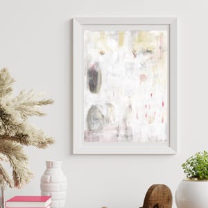 May include: A framed abstract wall art print featuring a white background with soft gold, grey, and muted pink accents. The composition includes vertical drips, faint text elements, and organic shapes, all enclosed within a clean white frame.