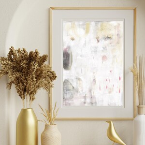 May include: A framed abstract wall art print featuring a white background with subtle grey, gold, and pink accents. The artwork displays soft, layered textures and faint vertical lines, enclosed in a light-colored wooden frame with a white mat border.