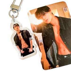 May include: A clear acrylic keychain with a silver clasp featuring a photo of a person in a black jacket. Beside it is a photo card with the number "01" visible.