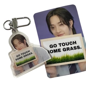 以下が含まれることがあります： 「GO TOUCH SOME GRASS」の文字が入った看板を持つ人物が写った、透明アクリルキーホルダーとフォトカード。看板には緑の草の縁取りがあります。キーホルダーにはシルバーの留め金が付いています。