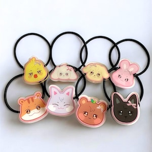 Pode incluir: Uma coleção de oito elásticos de cabelo pretos, cada um adornado com um rosto de animal de desenho animado diferente. Os rostos incluem um pato, um cão, um coelho, um gato, um urso e um tigre, em várias cores. O fundo é branco.