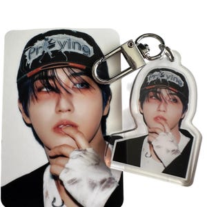 May include: A photograph and a keychain featuring the same image of a person wearing a black cap with the word "Praying" on it. The person is wearing a black blazer and has blue hair. The keychain has a silver clasp.