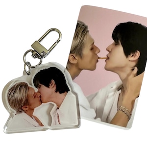 May include: A clear acrylic keychain with a silver clasp featuring a photo of two people kissing. A photo card shows two people sharing a snack. The background is a light pink color.