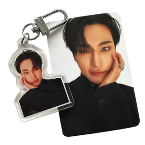 May include: A clear acrylic keychain and a photo card featuring a person with dark hair and a black outfit. The keychain has a silver clasp. The photo card and keychain have the same image of the person.