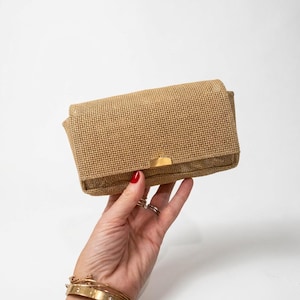 Può includere: Una pochette rettangolare realizzata in un materiale intrecciato color marrone chiaro. La borsa presenta un design a patta ripiegabile fissato con una chiusura rettangolare in metallo dorato liscio e lucido, centrata sul bordo anteriore.