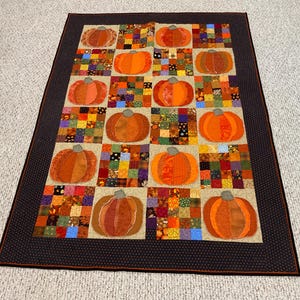 Op de afbeelding: Een handgemaakte quilt met een raster van oranje pompoenappliqués op een beige achtergrond. Elke pompoen staat tegen een patchwork vierkant van verschillende herfstthema stoffen. De quilt is omlijst door een donkerbruine rand met kleine witte stippen.