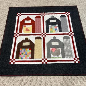 May include: A square quilt featuring four barn scenes with silos. The barns are red, black, and brown, with patchwork details and a sheep. The quilt has a white border with red and black accents and a dark gray border.