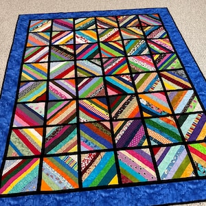 Puede incluir: Un edredón colorido con un diseño de patchwork. El edredón presenta una cuadrícula de cuadrados, cada uno lleno de rayas diagonales en varios colores, incluyendo rojo, naranja, amarillo, verde, azul y morado. Un borde azul rodea el edredón.