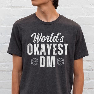 May include: A dark gray t-shirt with the text "World's Okayest DM" in white with two white dice on either side of the text.