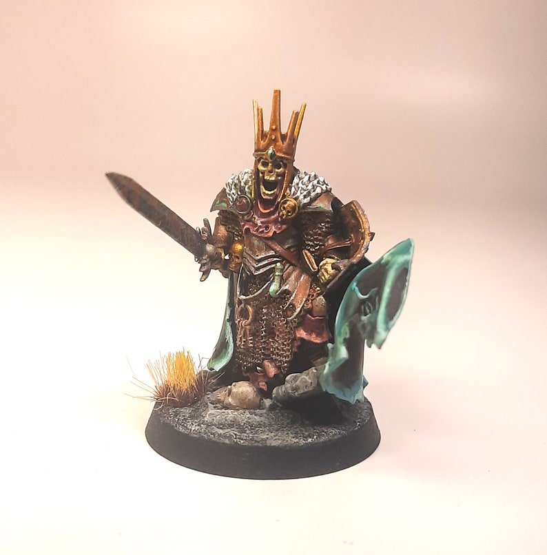 May include: A painted miniature of a skeletal figure wearing a golden crown and chainmail armour, holding a sword and a shield. The figure is standing on a black base with a small patch of brown grass.