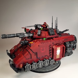May include: A red and black plastic model of a tank with a skull and crossbones on the side. The tank has a large cannon and a smaller machine gun on top. The tank is sitting on a grey base.
