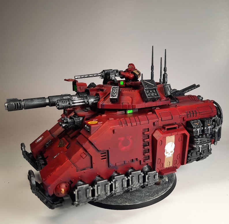 May include: A red and black plastic model of a tank with a skull and crossbones on the side. The tank has a large cannon and a smaller machine gun on top. The tank is sitting on a grey base.