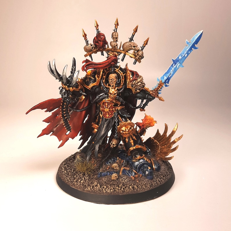 May include: A black and gold painted miniature figure of a warrior with a red cape and a blue sword. The figure is standing on a base with a fallen figure and skulls.