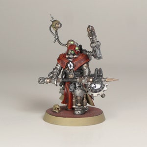 May include: A miniature metal figure of a robot wearing a red hooded cloak and holding a large metal weapon. The robot has a skull on its chest and green glowing eyes. The figure is standing on a round base.