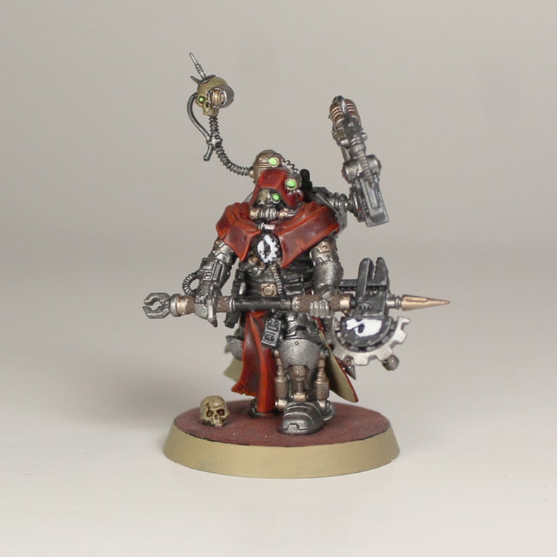 May include: A miniature metal figure of a robot wearing a red hooded cloak and holding a large metal weapon. The robot has a skull on its chest and green glowing eyes. The figure is standing on a round base.