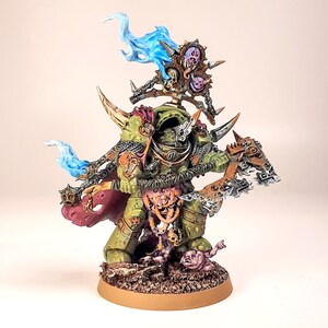 May include: A green and brown painted miniature figure of a Chaos Space Marine with a large axe and a skull-like helmet. The figure is standing on a brown base with a skull and bones. The figure is surrounded by blue and purple flames.