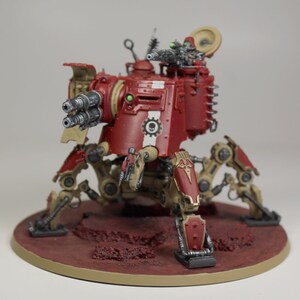 May include: A red and tan painted miniature war machine with a large gun and multiple legs. The machine is on a circular base with a red and tan painted surface.