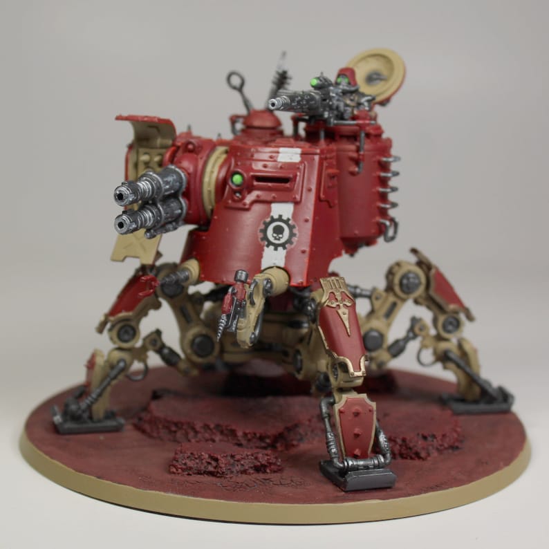 May include: A red and tan painted miniature war machine with a large gun and multiple legs. The machine is on a circular base with a red and tan painted surface.