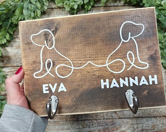 Può includere: Un cartello di legno con due sagome di cani e i nomi "Eva" e "Hannah" scritti sotto. Il cartello ha due ganci metallici attaccati alla parte inferiore.