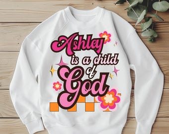 Può includere: Felpa bianca con la scritta "Ashley is a child of God" in rosa e nero. Il design include fiori rosa, quadrati arancioni e accenti a stella. Un top casual e confortevole.