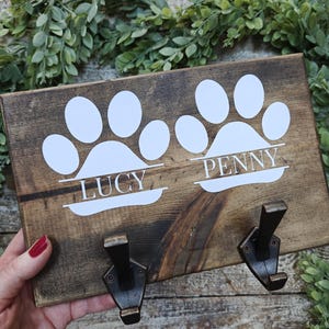May include: A wooden pet leash holder with two metal hooks. The holder features white paw prints and the names "LUCY" and "PENNY" in white lettering. The wood has a dark brown stain and is decorated with faux greenery.