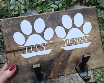 Può includere: Un porta guinzaglio per animali domestici in legno con due ganci in metallo. Il supporto presenta impronte di zampe bianche e i nomi "LUCY" e "PENNY" in lettere bianche. Il legno ha una macchia marrone scuro ed è decorato con finta vegetazione.
