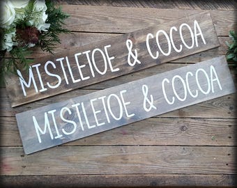 Può includere: Due cartelli rettangolari in legno con le parole "MISTLETOE & COCOA" in scrittura bianca. Un cartello è tinto di marrone scuro, l'altro di grigio chiaro. I cartelli sono esposti su una superficie di legno rustico.
