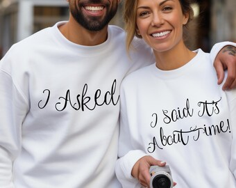 Può includere: Una coppia indossa felpe bianche con un set di testo abbinato. La felpa dell'uomo dice "I Asked" e la felpa della donna dice "I said It's About Time!"