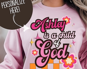 Può includere: Felpa rosa chiaro con la scritta "Ashley is a child of God" in stile retrò. Il design include fiori rosa, stelle e un motivo a scacchi arancione e bianco. Sono visibili anche le parole "Personalize Here!"