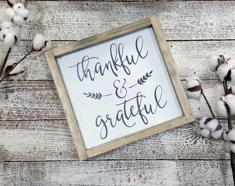 Può includere: Un cartello quadrato in legno con le parole "thankful & grateful" in corsivo blu scuro. Il cartello ha una cornice grigio chiaro ed è posto su uno sfondo di legno imbiancato. Steli di cotone sono disposti attorno al cartello.