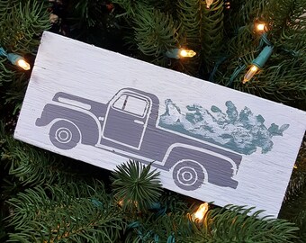 Può includere: Un cartello rettangolare in legno con un camioncino vintage grigio che trasporta un albero di Natale verde. Il camion è dipinto su uno sfondo bianco. Il cartello è circondato da aghi di pino verdi e luci natalizie.