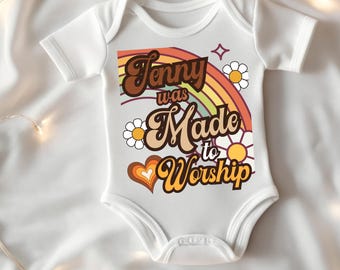 Può includere: Body da bambino bianco con maniche corte. Il davanti presenta un design retrò con un arcobaleno, margherite e la scritta "Jenny was Made to Worship" in lettere marroni e arancioni. È inclusa anche una grafica a forma di cuore.