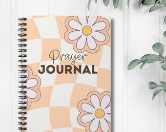 Può includere: Un diario di preghiera a spirale con una copertina a scacchi color pesca e bianco. La copertina presenta due margherite bianche con centri gialli. Il testo "Prayer Journal" è stampato in marrone sulla copertina.