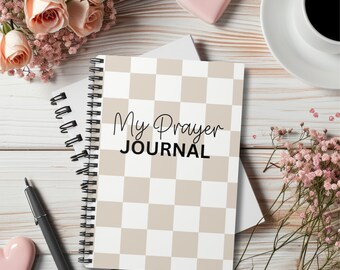 Può includere: Un diario a quadri bianchi e beige con il testo "My Prayer JOURNAL" in lettere nere. Il diario è su una superficie di legno bianco con fiori rosa e un cuore rosa.