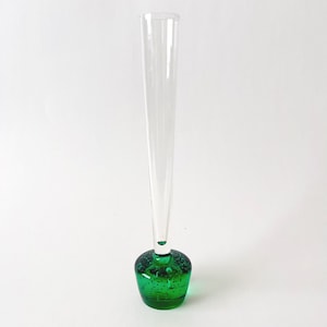May include: A clear glass bud vase with a long, slender neck and a rounded, emerald green base. The base has a bubbly texture. The vase is designed to hold a single flower or small floral arrangement.