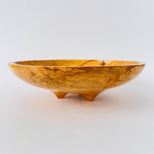 May include: A wooden bowl with a natural wood finish and three legs. The bowl is made of a light brown wood with darker brown streaks.