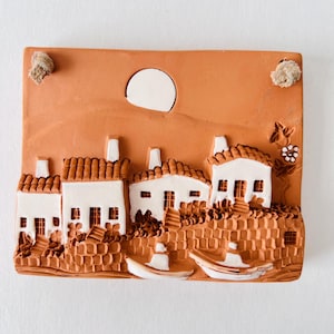 May include: A terracotta wall plaque depicting a small village with white houses, red tile roofs, and a full moon in the sky. The houses are clustered together and have small windows and doors. The plaque is a 3D relief sculpture with a textured surface.