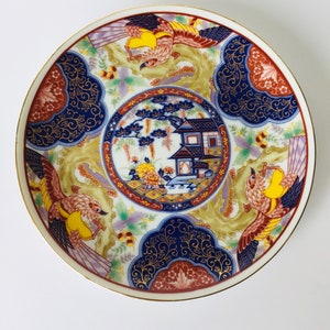 May include: A white ceramic plate with a gold rim features a colorful Japanese-inspired design. The design includes a central image of a house with a tree, surrounded by stylized birds and floral patterns in shades of blue, red, yellow, and gold.