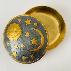 Könnte beinhalten: Eine Messing-Schmuckschatulle mit einem Deckel, der eine Sichelmond und Sterne auf einem dunkelgrauen Hintergrund zeigt. Die Sonne ist in der Mitte des Deckels mit einem Gesicht dargestellt. Der Boden der Schachtel ist aus einfachem Messing.