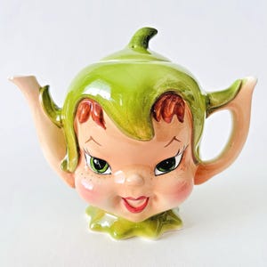 May include: A charming ceramic teapot in the shape of a smiling face, featuring green hair and a matching lid. The face is peach-toned with red accents, green eyes, and a red smiling mouth. The teapot has a spout and handle, set against a white backdrop.