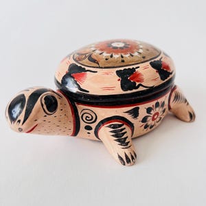 May include: Hand-painted ceramic turtle trinket box with a hinged lid. The turtle is decorated with black, red, and brown floral patterns.
