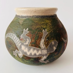 May include: A small, round ceramic pot featuring a textured, mottled green and brown surface with a prominent light grey lizard silhouette. The vessel has a smooth, cream-coloured rim with fine crackle details and dark horizontal line accents.