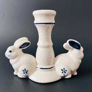 May include: White ceramic candle holder with two rabbit figurines on either side. The holder has a crackled glaze and blue accents.