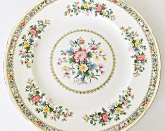 May include: A white ceramic plate with a floral design. The plate features a central floral bouquet and a border of flowers in pink, yellow, and blue. The rim has a gold trim.