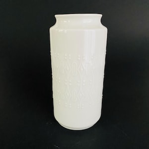 Könnte beinhalten: Eine weiße Keramikvase mit zylindrischem Körper und leicht ausgestelltem Rand. Die Vase weist ein sich wiederholendes Muster aus erhabenen, stilisierten floralen oder geometrischen Designs auf. Der Hintergrund ist einfarbig schwarz.