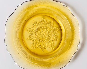 May include: A round, translucent amber-colored glass plate featuring a central embossed star-like pattern surrounded by a circular border. The rim of the plate has a scalloped edge with a delicate, repeating etched design along the outer border.