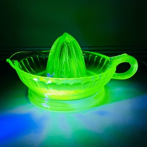May include: A vintage, bright green glass juicer with a conical reamer and a circular bowl with a handle. The juicer is made of a translucent glass, and glows under a black light. The bowl has a decorative pattern. The juicer is likely made of uranium glass.